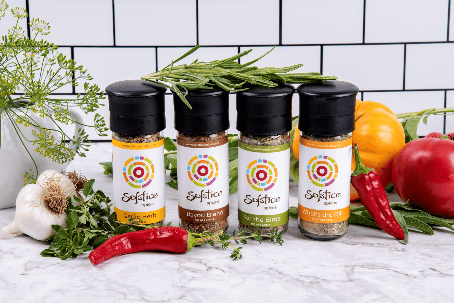 Four glass jars of Solstice Spices seasoning with black grinder caps surrounded by red chile peppers, garlic, fresh herbs, and yellow and red heirloom tomatoes. The background is white kitchen tile with black grout.