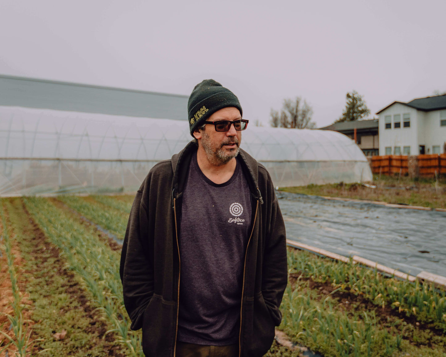 The owner of Solstice Spices stands in front of several rows of garlic growing under a grey sky with high tunnels in the background.