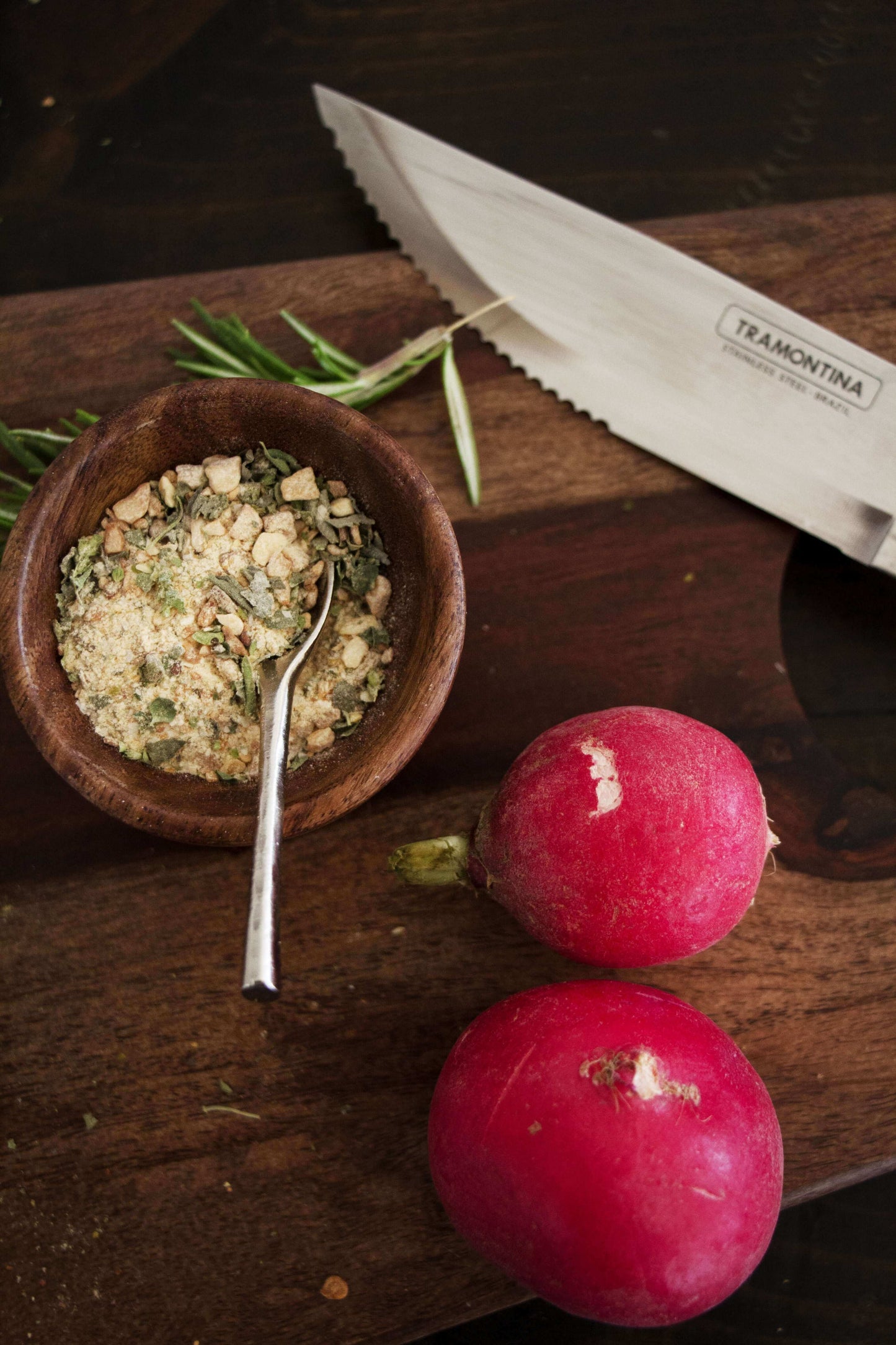 Overhead photo of a wooden cutting board with a small wooden bowl of dried spices on top and two pink radishes with the blade of a knife.