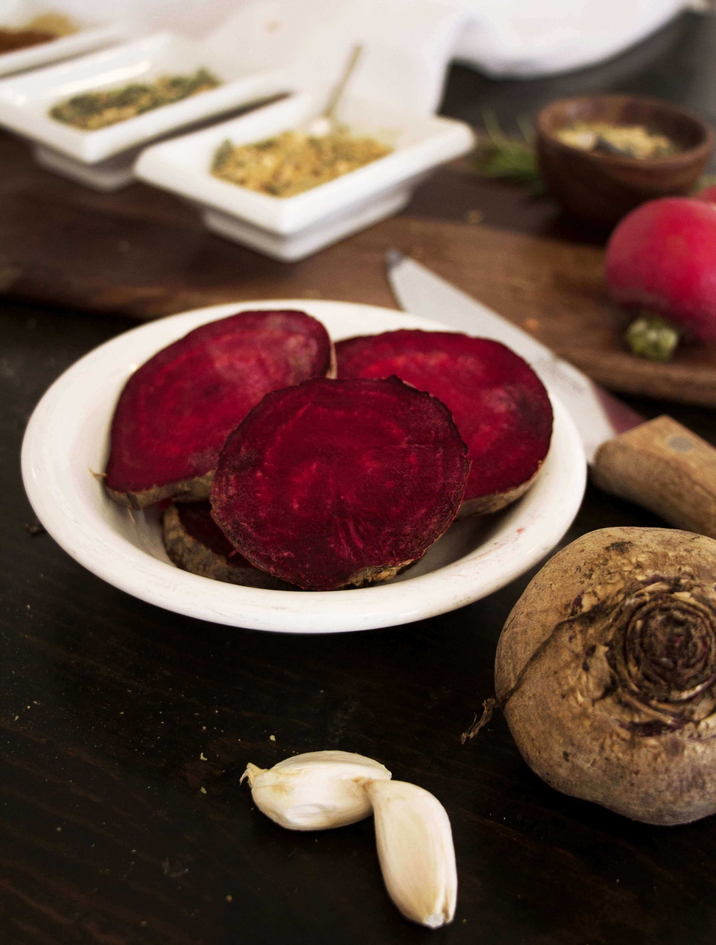 Sliced beetroot in a white ceramic bowl with two cloves of garlic in front and a whole beet on the side. In the background there is a knife and small white bowls of spices.