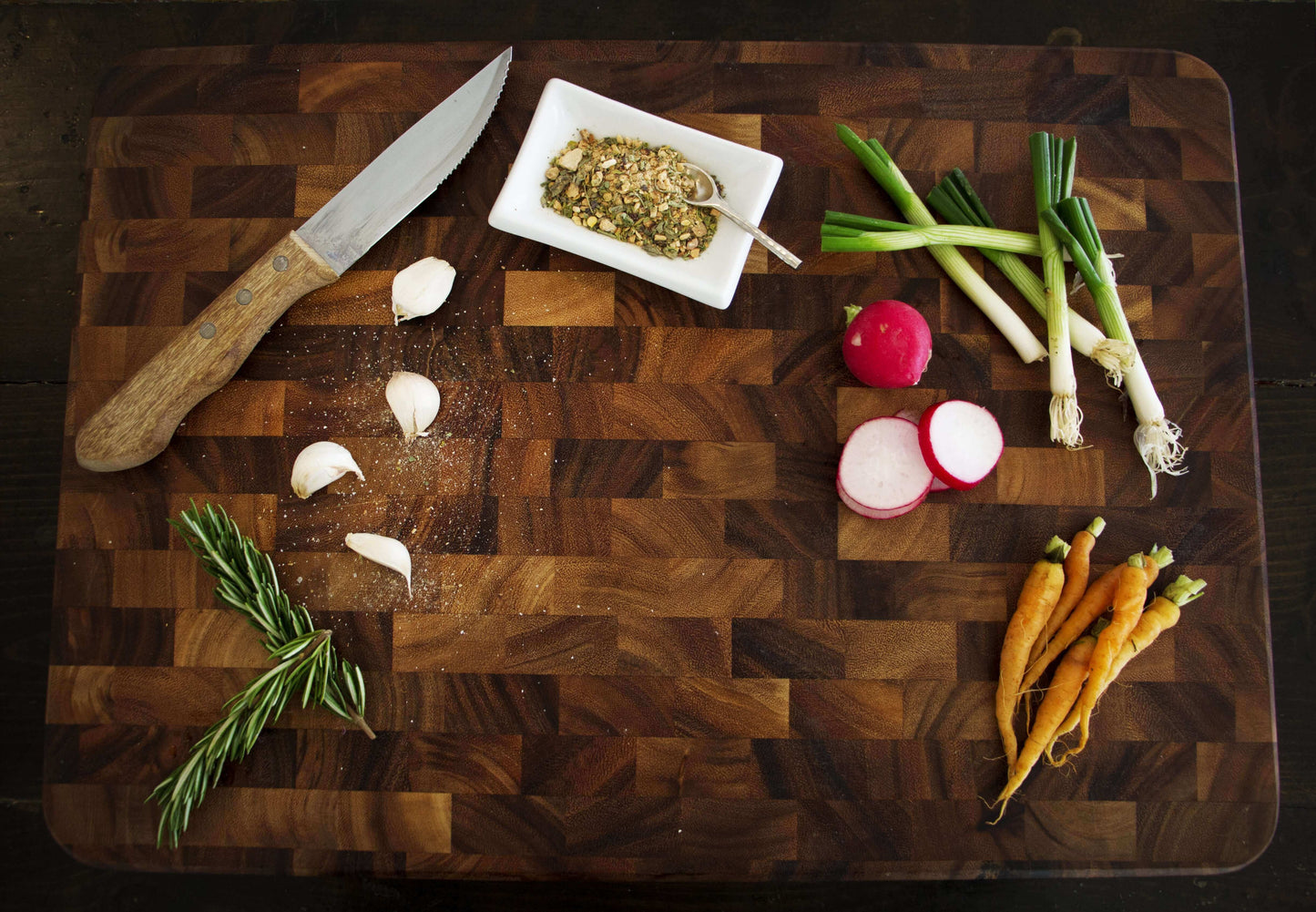A wooden cutting board with a knife and rosemary, garlic, green onions, radishes, and carrots.