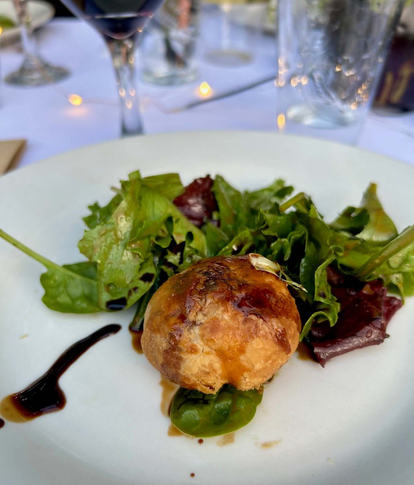 Perfectly browned puff pastry with a balsamic glaze and mixed greens on a white plate with a glass of wine in the background.