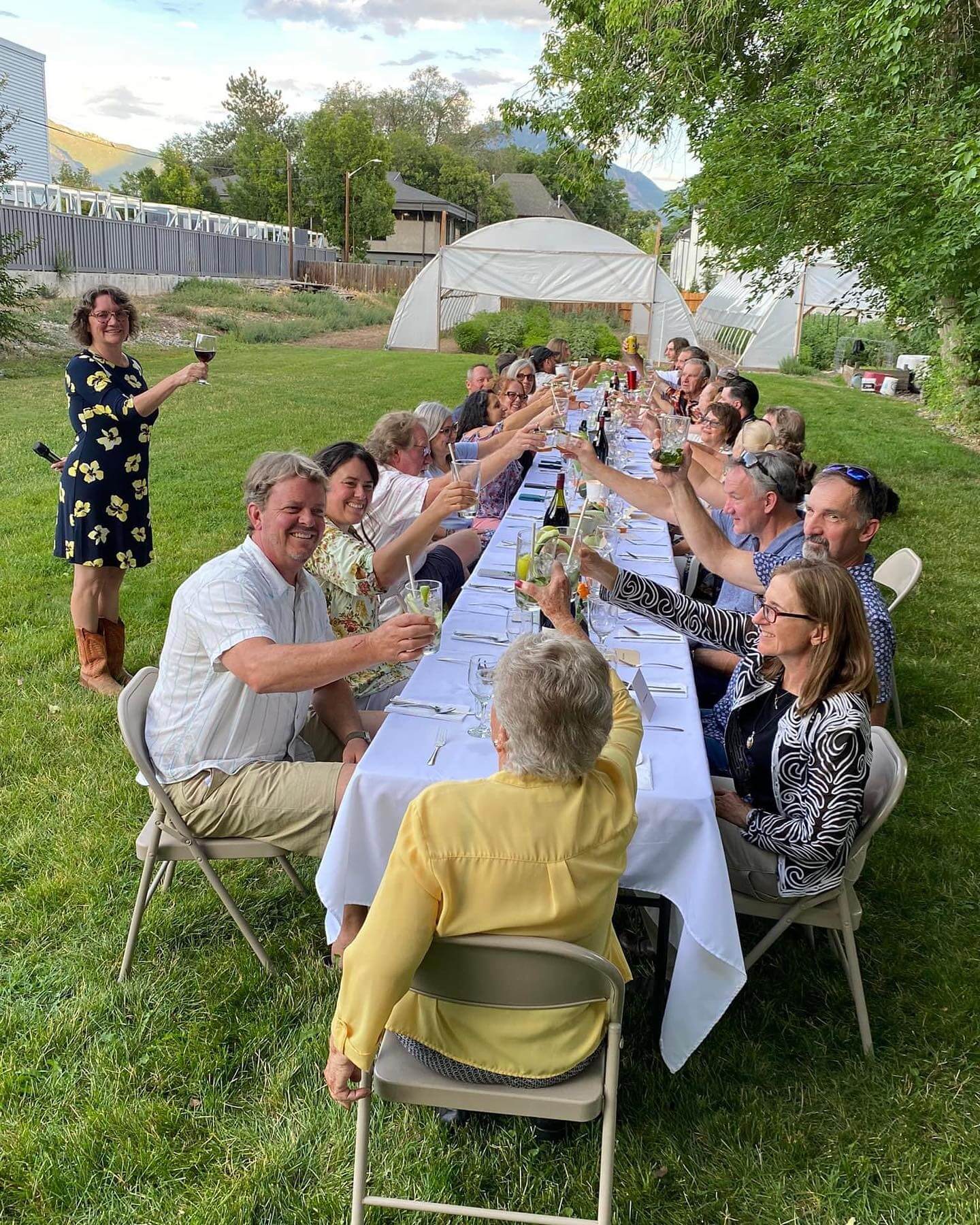 Dining guests enjoying a toast seated at a long table with a white tablecloth and farm high tunnels in the background.