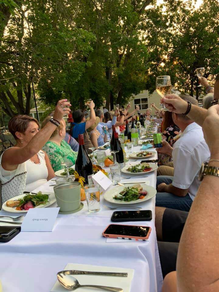 Dinner guests enjoying a toast while seated around a long table with a white tablecloth and trees in the background.