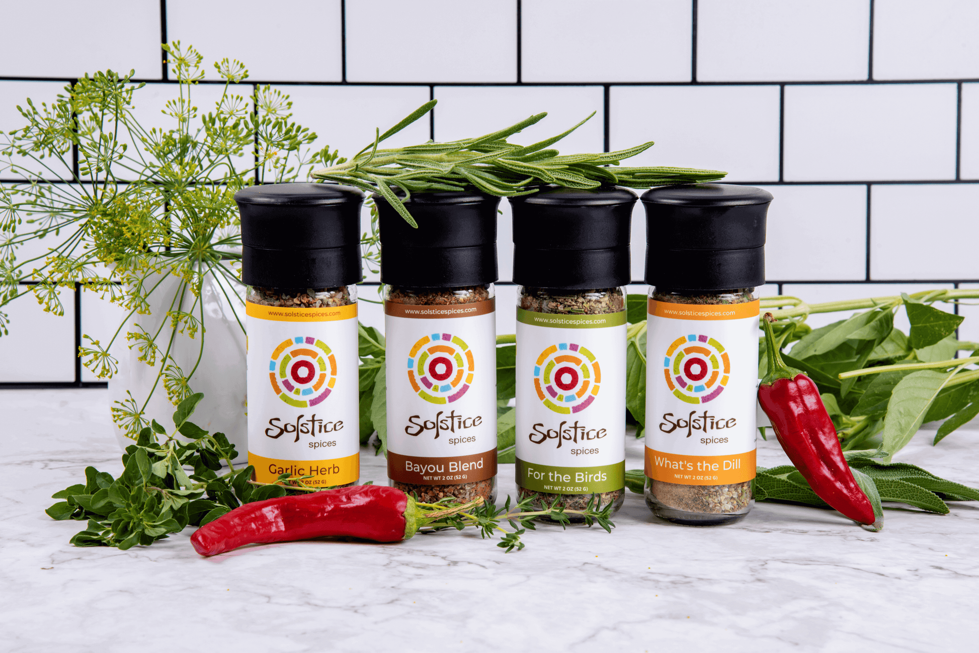 4 glass Solstice Spices grinder jars surrounded by fresh herbs and red chile peppers. The backdrop is white kitchen tiles with black grout.