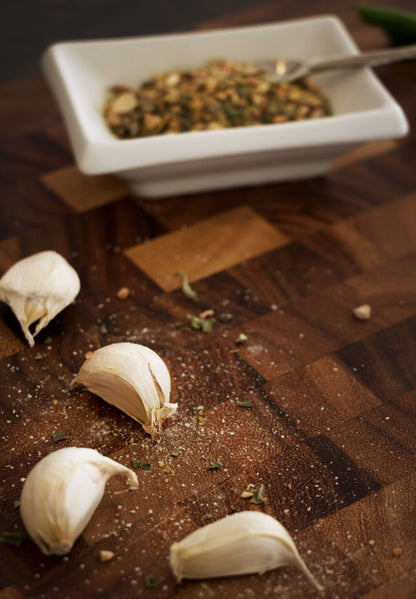 Four cloves of fresh garlic on a wooden cutting board with a white ceramic dish of dried spices in the background.