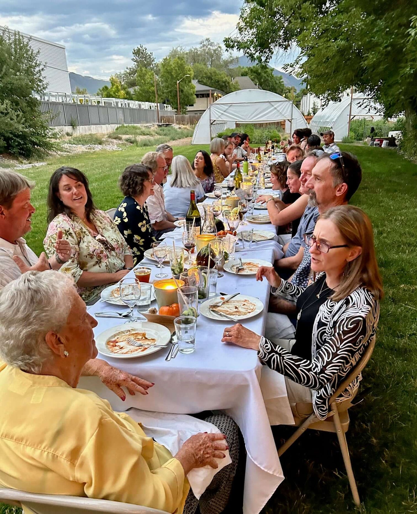 People enjoying dinner seated at a long table with a white tablecloth and farm tunnels in the background.