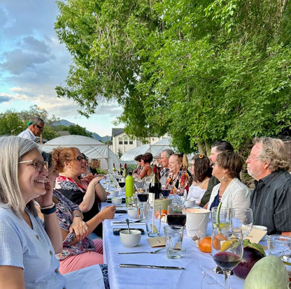 People enjoying dinner at a long table covered in a white tablecloth and trees and farm high tunnels in the background.