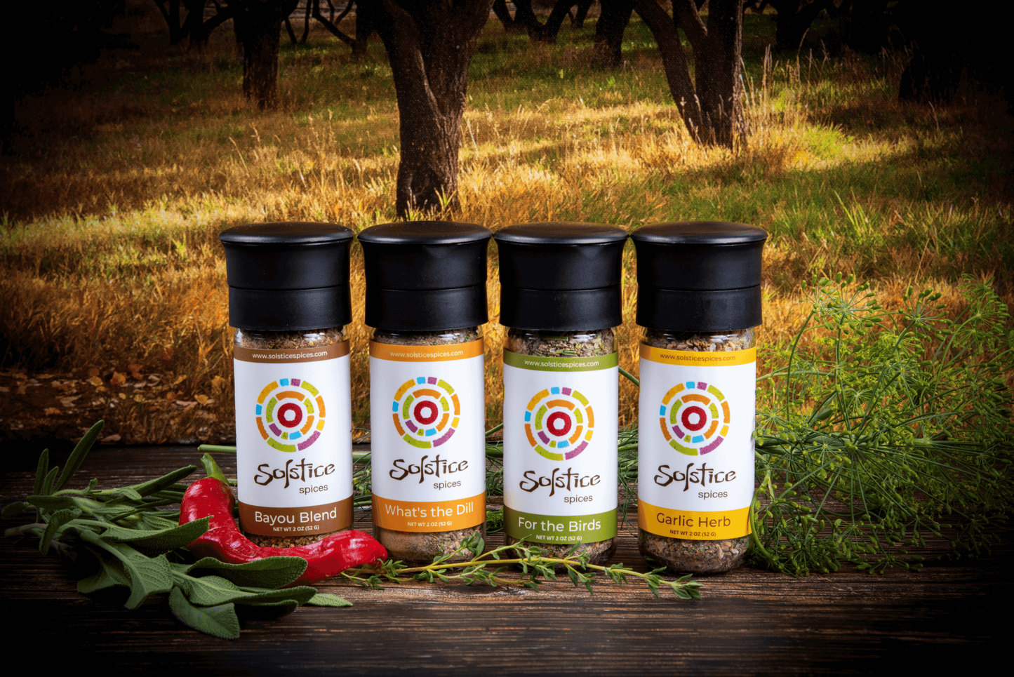 Four glass spice grinder jars with Solstice Spices labels. The jar on the far left is Bayou Blend, next over is What's the Dill, next is For the Birds, and the far right is Garlic herb. Fresh herbs and a red chile pepper surround the spice jars and the background is a yellow field with fruit trees.