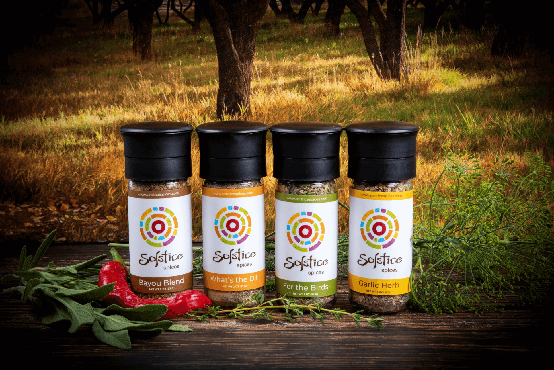 Four glass spice grinder jars with Solstice Spices labels. The jar on the far left is Bayou Blend, next over is What's the Dill, next is For the Birds, and the far right is Garlic herb. Fresh herbs and a red chile pepper surround the spice jars and the background is a yellow field with fruit trees.