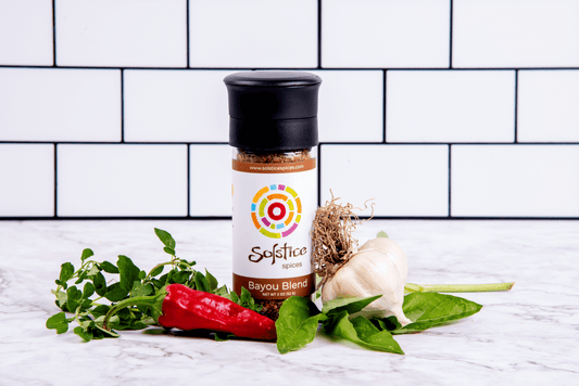 A glass spice grinder jar with a Solstice Spices label that has a brown boarder with the words "Bayou Blend" in white. The jar is surrounded by a fresh bulb of garlic, a red chile pepper, and fresh herbs. The background is white kitchen tiles with black grout.