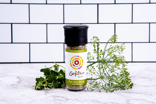 A glass spice grinder jar with a Solstice Spices label that has a green boarder and the words "For the Birds" in white. Surrounding the jar are fresh cut herbs and in the background are white kitchen tiles with black grout.