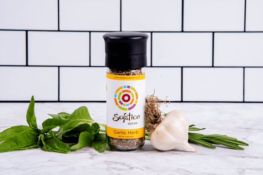 A glass spice grinder jar with a Solstice Spices label that has a yellow boarder and the words "Garlic Herb" in brown letters. Surrounding the jar is a farm fresh bulb of garlic and basil leaves. In the background there are white kitchen tiles with black grout.