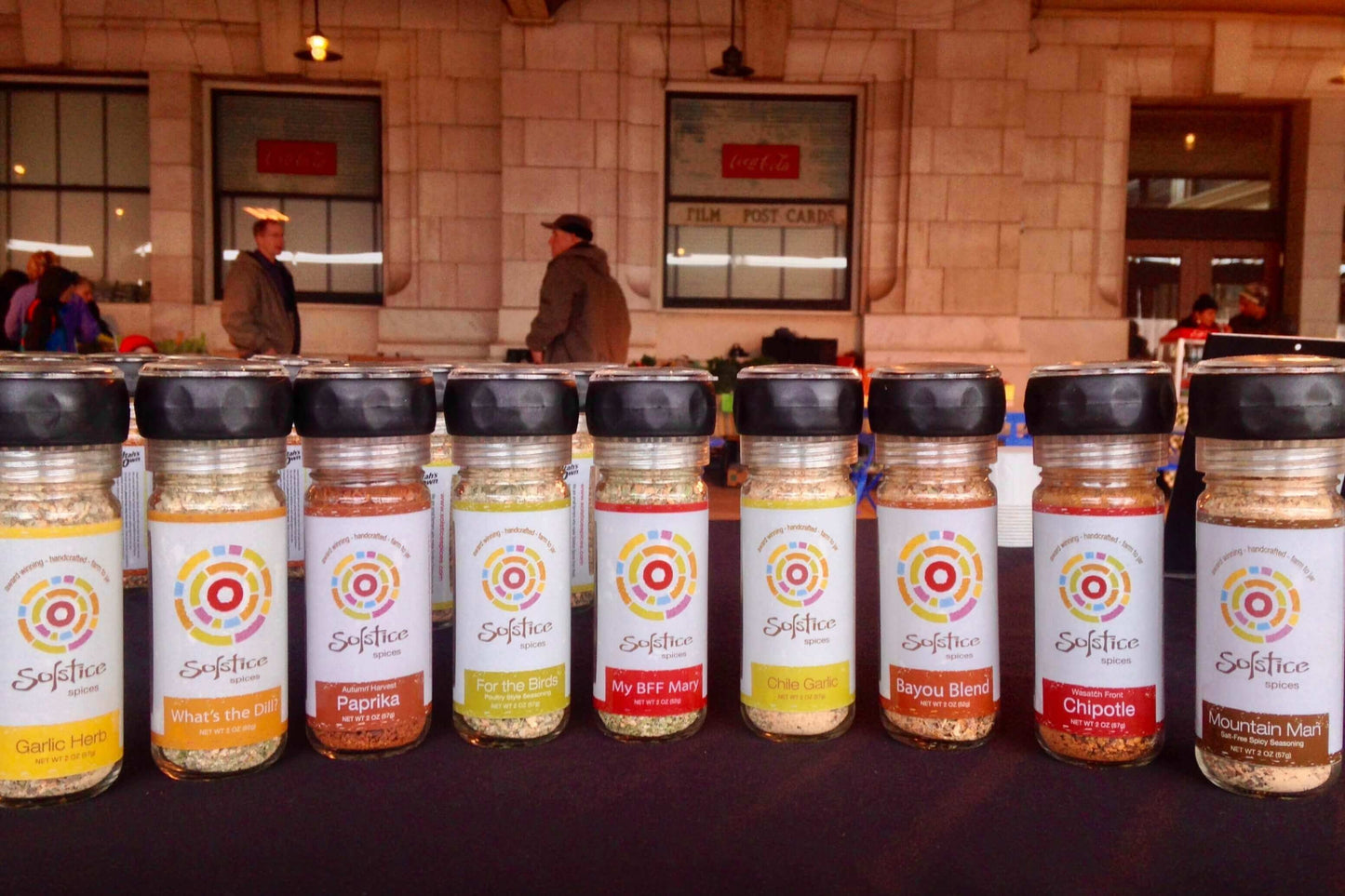 10 different flavors of Solstcie Spices glass grinder jars in a row with the Salt Lake Downtown Farmer's Market at the historic Rio Grande building in the background