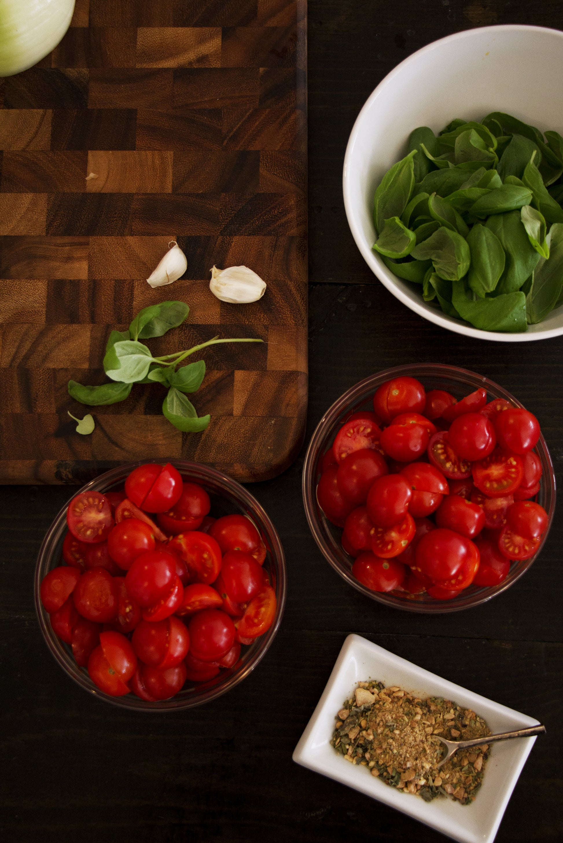 Overhead shot of chopped cherry tomatoes in two small wooden bowls with a white bowl of herbs, a small rectangular dish of dried spices, and a wooden cutting board.