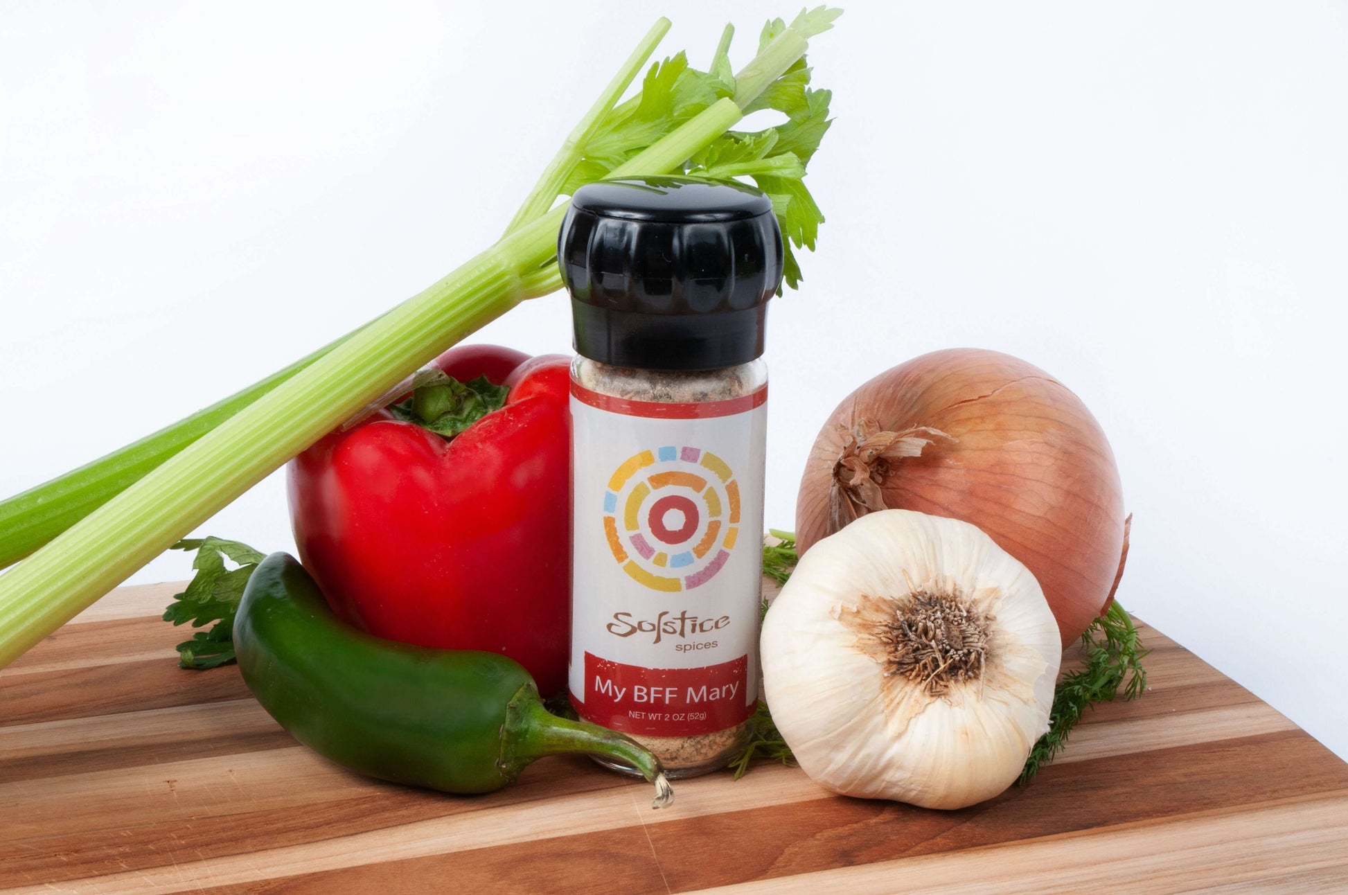 A jar of Solstice Spices My BFF Mary Seasoning surrounded by a stalk of fresh celery, a red bell pepper, a bulb of garlic, a yellow onion, and a green jalapeno pepper all on top of a wooden cutting board with a white background.