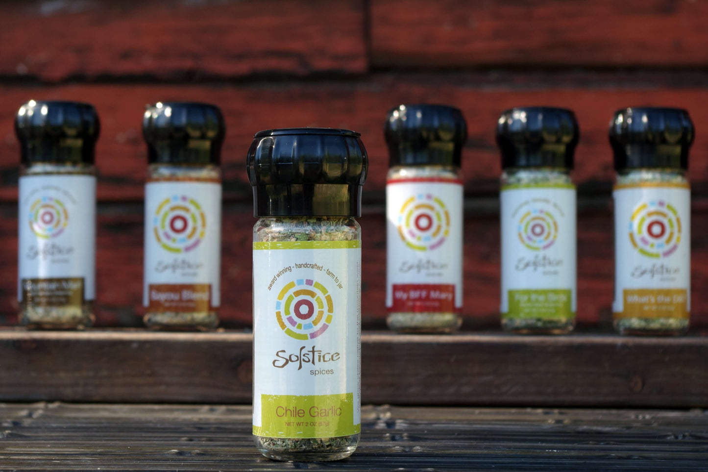 Solstice Spices Chile Garlic Seasoning in front of 5 other jars of spices in front of a red wooden barn at a farm.