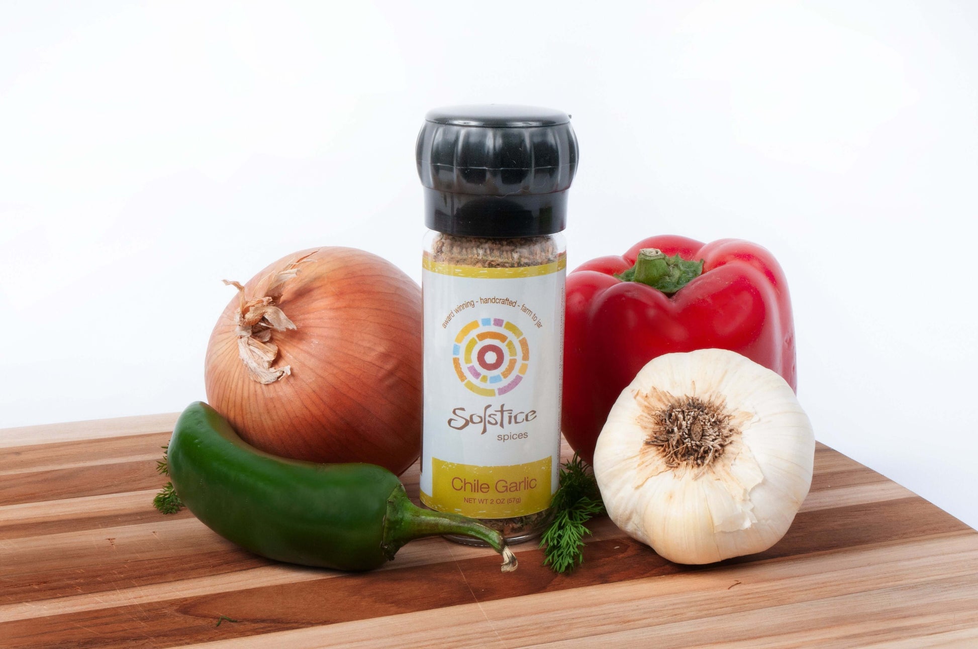 A Solstice Spices glass grinder jar on a wooden cutting board surrounded by a yellow onion, garlic bulb, red bell pepper, and green jalapeno.