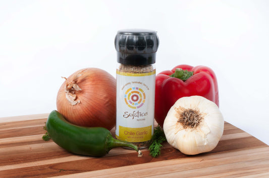 A Solstice Spices glass grinder jar on a wooden cutting board surrounded by a yellow onion, garlic bulb, red bell pepper, and green jalapeno.