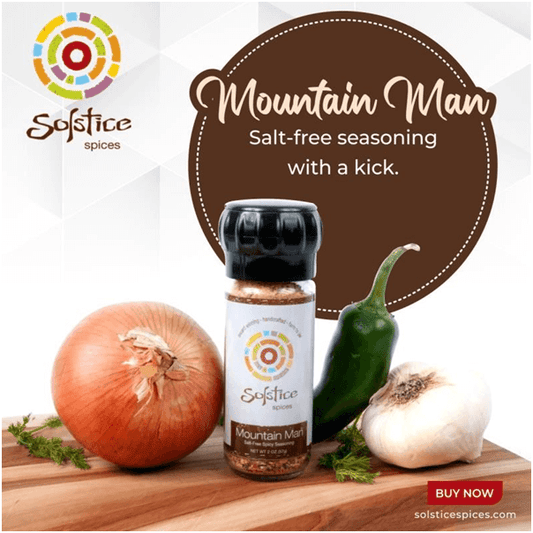 A jar of Solstice Spices Mountain Man seasoning surrounded by a whole onion, garlic bulb, and a green jalapeno pepper. In the background is a brown circle with the words "Mountain Man Salt-free seasoning with a kick." and the Solstice Spices logo.